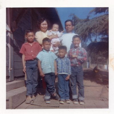 A photograph of the Reyes family at the labor camp of Reiter Berries off of San Andreas Rd in Watsonville. From left to right, in the red shirt is Teddy Reyes, Raymond Reyes, Roland Reyes, and Joe Reyes (seen sticking his tongue out). In the back row is mother and father, Tecla and Jose Reyes with their baby, Elizabeth Reyes.