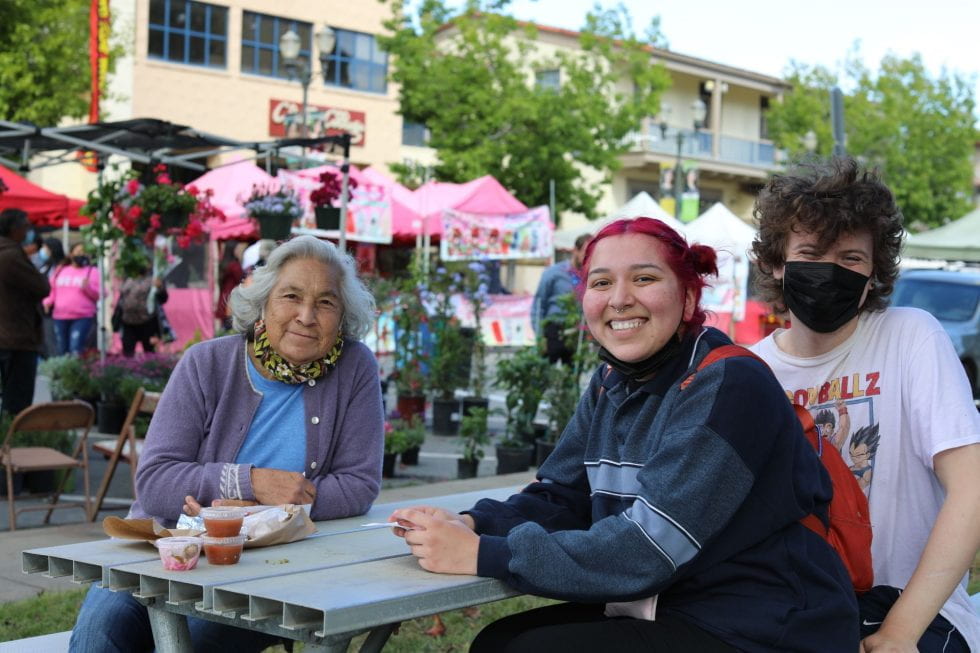 two students sitting with an older woman at a picnic table with leftovers, smiling at the camera