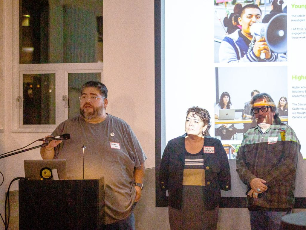 three participants in front of projector screen, one at podium, two waiting to the right